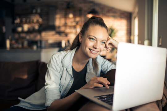 Young woman in casual clothing using laptop and smiling while working indoors