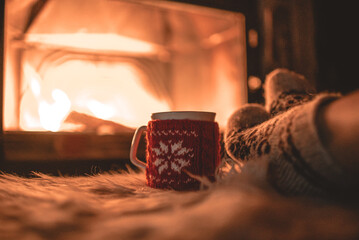 Feet in woollen socks by the Christmas fireplace. Woman relaxes by warm fire with a cup of hot drink and warming up her feet in woollen socks. Close up on feet. Winter and Christmas holidays concept.