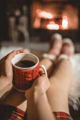 Woman holding a cup of tea by the Christmas fireplace. Woman relaxes by warm fire with a cup of hot drink and warming up her feet in woollen socks. Close up on feet. Winter, Christmas holidays concept