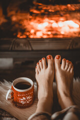 Bare woman feet by the cozy fireplace. Woman relaxes by warm fire with a cup of hot drink and warming up her feet. Close up on feet. Winter and Christmas holidays concept.