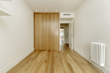 Empty bedroom with a custom-made built-in wardrobe with light oak wooden doors, a white radiator on the wall and air conditioning through ducts in the ceiling and floor of the same material.