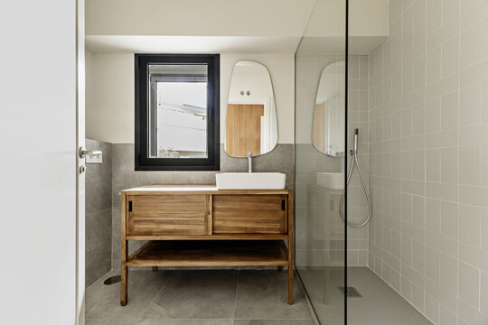 Bathroom in an apartment with wooden furniture with sliding doors, floating white porcelain sink, mirror hanging on the wall and shower cabin with armored glass screen