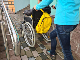 Adult woman mother helps daughter child in wheelchair climb ramp in city