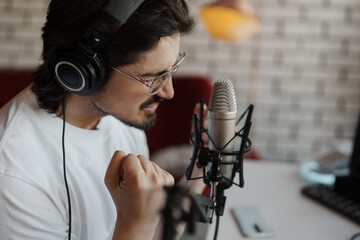 Side view of passionate male singer in headphones sitting at table near microphone and recording...