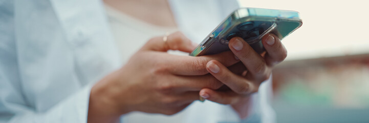 Closeup of young woman holds smartphone in her hands and scrolls through the news feed. Close-up of girl hand uses mobile phone outdoors - obrazy, fototapety, plakaty