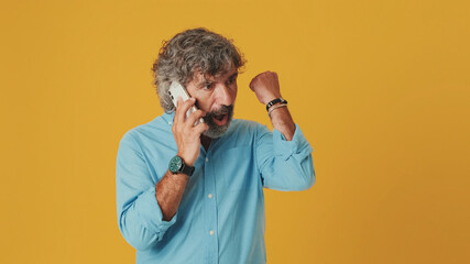 Surprised elderly grey-haired bearded man wears blue shirt, talking on mobile phone, found out great big win, celebrating victory, isolated on orange background in studio