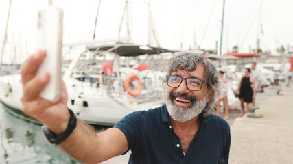 Close up, happy middle-aged man in glasses sits in the port on yacht background uses smartphone,...
