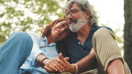 Fototapeta premium Happy retired couple sitting in the park holding hands and smiling resting on the grass in the park in autumn