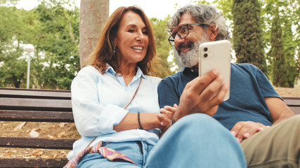 Retired couple talking, having fun using mobile phone while sitting outside in the park in autumn