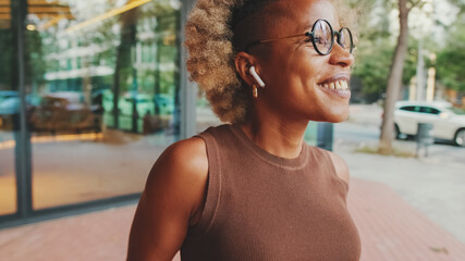 Happy young woman in glasses wearing brown top in wireless headphones dancing outdoors in city...