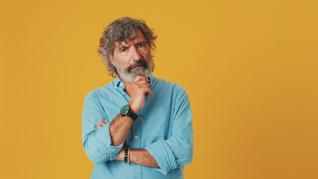 Elderly gray-haired bearded man wears a blue shirt, looks at the camera listens carefully and agrees, isolated on an orange background in the studio