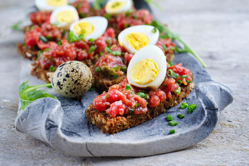 Vollkornbrot Schnitte mit Tartar Belag serviert mit Wachteleiern Kapern und Kräuter als Nahaufnahme auf einem schwarzen Design Tablett 