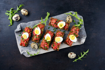 Vollkornbrot Schnitte mit Tartar Belag serviert mit Wachteleiern Kapern und Kräuter als Draufsicht auf einem schwarzen Design Tablett 