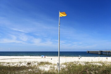 Yellow flag flapping in the wind atop a tall metal pole situated next to a tranquil blue sea