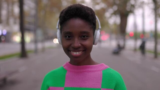 Portrait of young adult african woman in headphones looking at camera standing on street. Smiling african american female generation z hipster smiling over urban background.