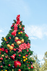A tall fir tree decorated with glass balls, toys, ornaments, Christmas lights, glitter, red polka dot bows,  bobbles, and baubles outdoors under blue sky. There's a red ribbon looped at the top.