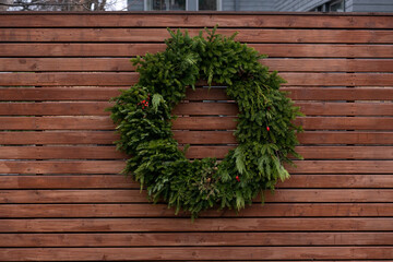 Fototapeta na wymiar Thick lush green branches in a circular shape with small red berries on a brown fence. The Christmas wreath has natural green fir and cedar boughs. The wall is made of narrow wooden brown boards. 
