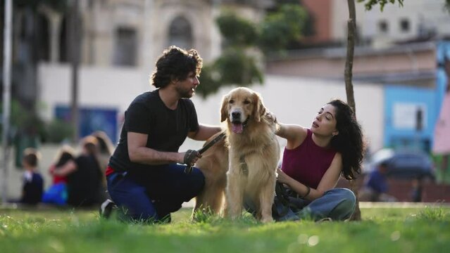 Candid couple sitting at park with their Dog on a leash. Young man and woman millennials enjoying sunny day outdoors with their Golden Retriever Dog Pet. Canine Companion concept