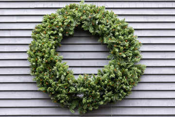Lush green fir branches in a circular shape on a grey wall. The Christmas wreath has natural green boughs. The wall is made of narrow wooden silver-gray boards.  There's sparkle on the tips of the fir