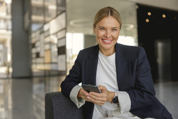 Cheerful businesswoman use phone sitting on modern office background and looks camera
