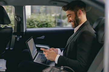 Handsome businessman in suit using personal computer and using mobile phone in taxi