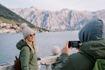 Husband photographs his wife leaning on the railings on the seashore overlooking the mountains. Back view