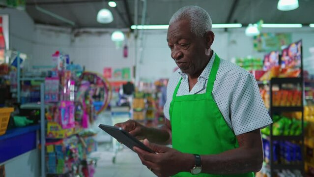 African American senior employee of supermarket using tablet device standing inside grocery store, job occupation of a black older person at workplace, checking inventory