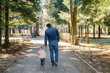 Dad leads a little girl by the hand through a sunny spring park. Back view