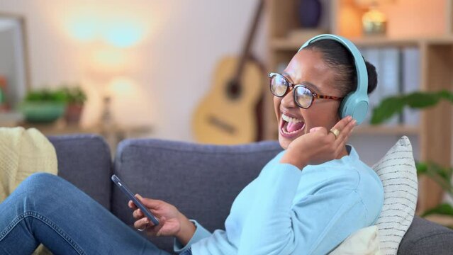 Woman streaming music on phone while wearing headphones and singing along while relaxing at home. Cheerful african female on couch listening to her favorite songs on a mobile app over the weekend