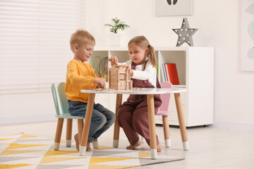 Little boy and girl playing with wooden house at white table indoors. Children's toys