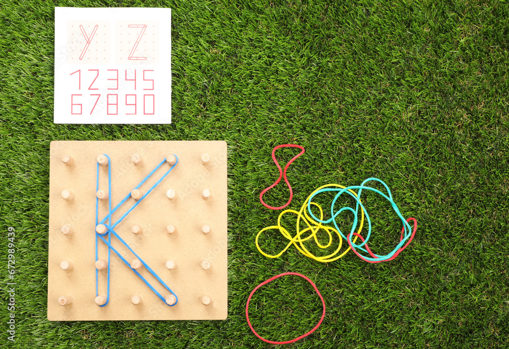 Wall mural Wooden geoboard with letter K made of rubber bands and activity book on artificial grass, flat lay. Educational toy for motor skills development