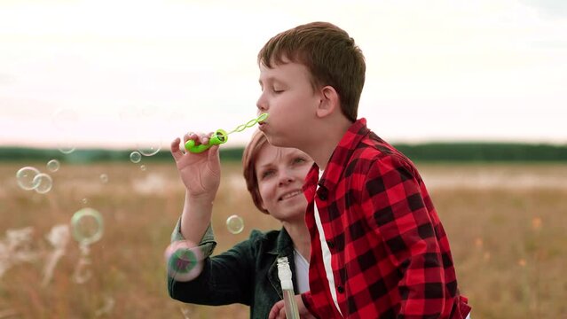 Cheerful boy in checkered shirt blows bubbles while mother helps on mowed field