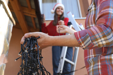 Woman in Santa hat and man decorating house with Christmas lights outdoors, selective focus