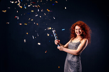 Red-haired, young woman in party laughing while blowing up party popper isolated over black wall. New year's concept, celebration birthday. copy space