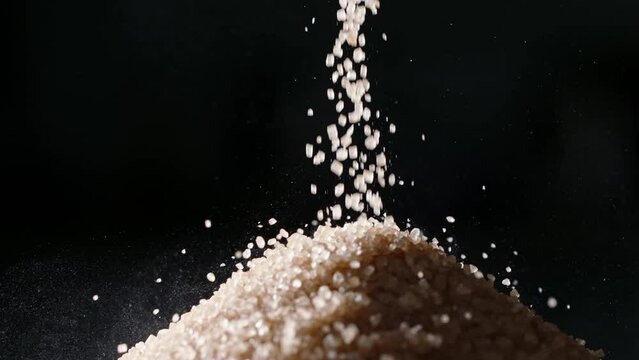 Brown cane sugar is poured into pile. Close up slow motion black background. Restricting sugar intake helps minimize risk of obesity, diabetes, and various other diseases that afflict many today. Diet