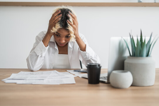 Broke,Frustrated african american girl holding her head sitting in front of pile of bills and invoices.Expenses,cost of living,debt concept.