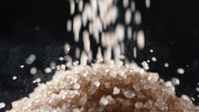 Brown cane sugar is poured into pile. Macro shot slow motion black background. Excessive in sugar is harbinger precursor of obesity and diabetes, necessitating its restriction to protect well-being