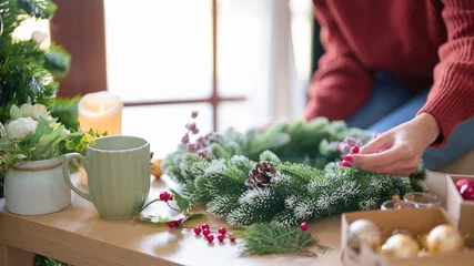 Fotobehang Young asian woman in sweater making christmas wreath and decorations with fir pine branch and red berries while prepare ornaments to celebrate for christmas festive holiday and winter seasons at home © snowing12