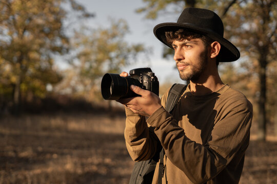 Portrait of travel photographer wearing hat with digital camera making photo of the nature at sunset.