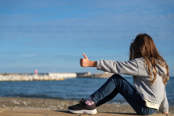 Niña en la playa, vista de espaldas, octubre, pelo largo suelto, jugando en la arena, sentada mirando al mar, mano en alto signo ok