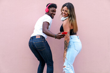 Cheerful African guy and Latina girl enjoy music on phone, dancing by pink wall.