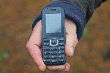 one old push-button mobile cell phone, black, plastic, small, compact, for calls and the Internet in the hand of a man on the street in the forest during the day