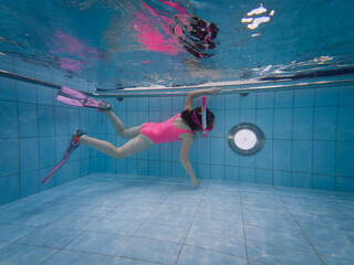 A little girl in a pink swimsuit is training to swim underwater with a snorkel and snorkel mask.