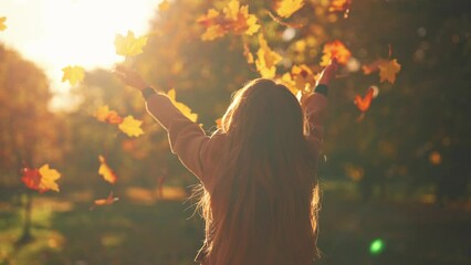 Young woman having positive good mood throwing fallen maple leaves in air in sunny autumn weather in park, back view. Outdoors recreation entertainment in fall season, playing with foliage concept.