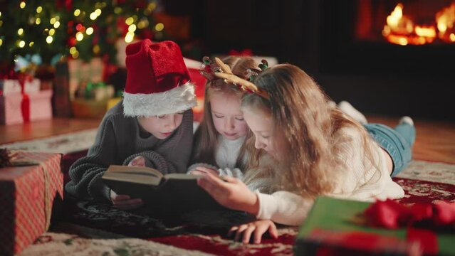 Children siblings reading book in Christmas holidays lying on floor near fireplace, Christmas tree in living ror om at home. Interested sisters brother look in book. Family atmosphere New year Xmas.