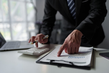 Young professional Asian businessman using laptop, computer and using phone. View financial statistics displayed in documents and work on a laptop computer at the company.