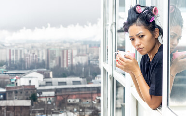 A young woman in curlers looks out of the window of a high-rise building in an industrial zone