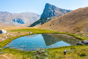 A hiker standing by a lake with grass around it and looking at a peak reflected in the water