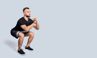 Serious handsome strong millennial european guy with beard in sportswear squat with fitness elastic band