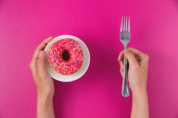 Female hands hold a donut on a plate and a fork on a pink background. Top view, flat lay. Sweet,...